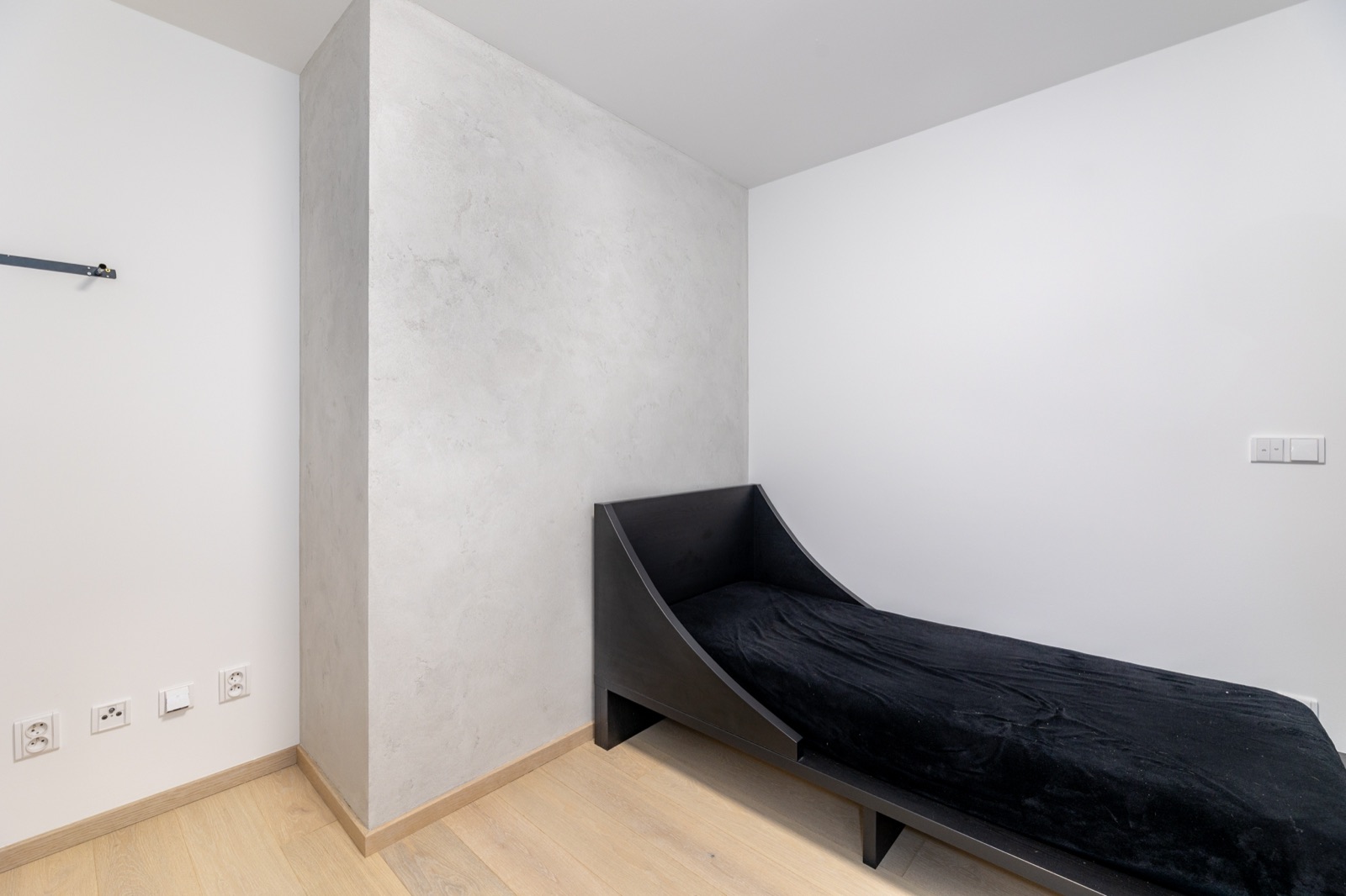 Minimalistická ložnice se šedou betonovou stěrkou v rohu a černým lehátkem — realizace Dehner